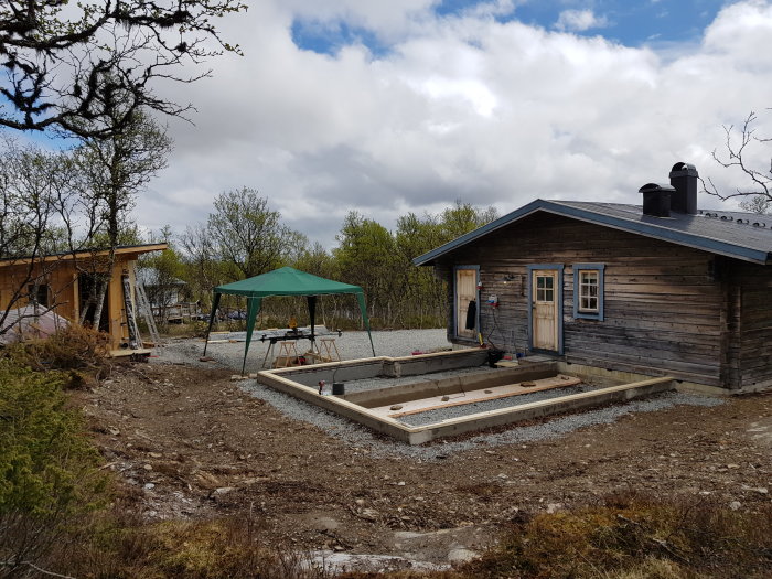 Pågående byggprojekt med en nyligen fastbultad syll och några väggreglar på plats, intill en befintlig bastu.