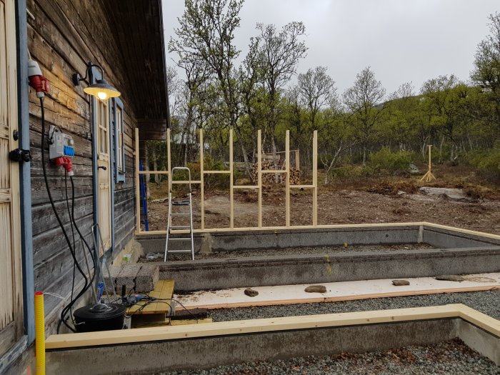 Byggplats med fastbultad syll och uppförda väggreglar vid ett bygge i dagsljus, med en befintlig byggnad intill.