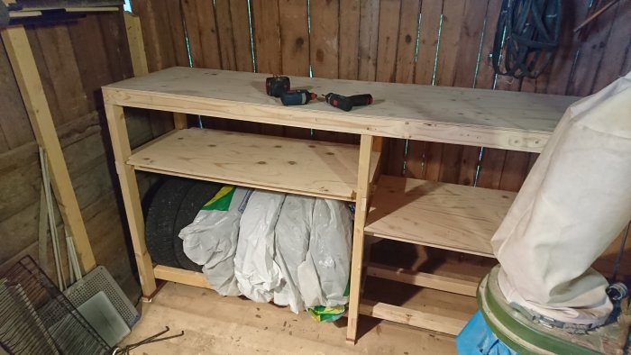 Nybyggd trähylla i verkstad med slipmaskin och kapad plywood, innebär framsteg i projektet.