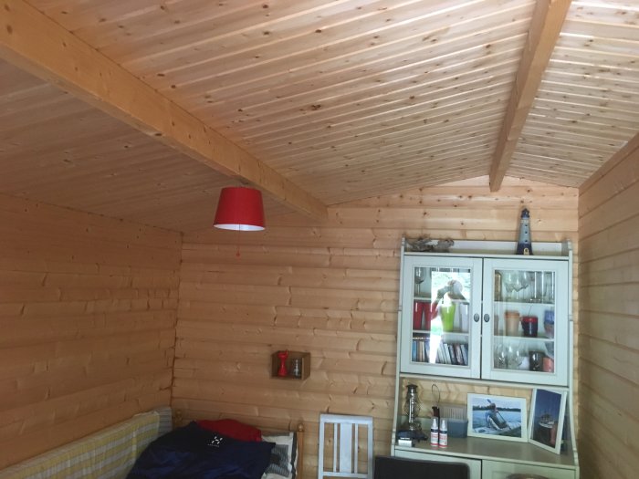 Inredning av en timrad friggebod med synliga takbjälkar, oisolerad vägg och tak, och en röd lampa.