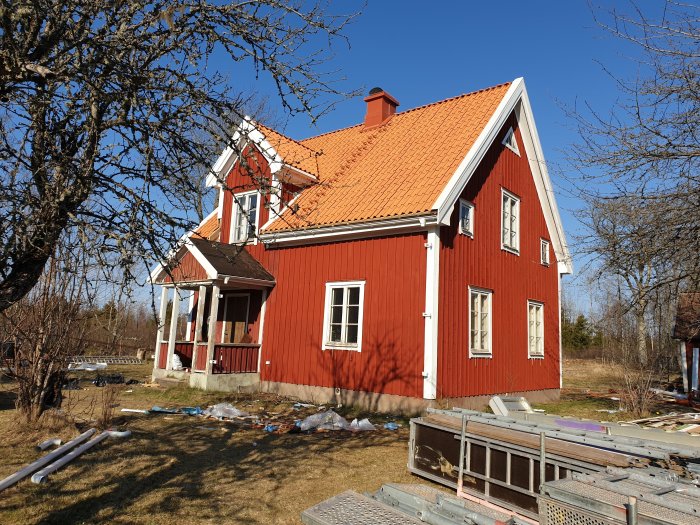 Nyrenoverat rött hus med nytt tegeltak, byggmaterial synligt på tomten under en solig dag.