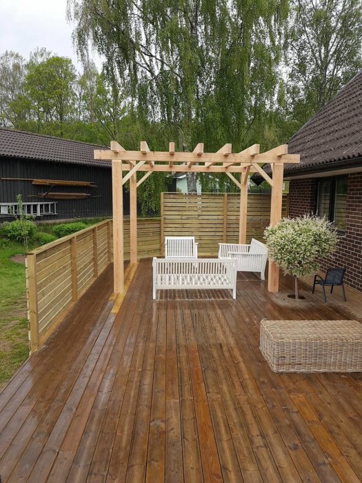 En nyligen byggd pergola på ett trädäck med sittmöbler i en trädgård.