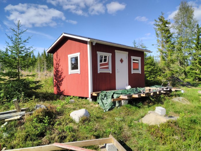 Rött trähus med lägre placerade vita fönster än dörren i en skogsglänta.