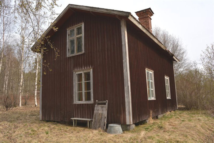 Rödfärgad timmerstuga med vitmålade knutar på en naturtomt, omgiven av träd.