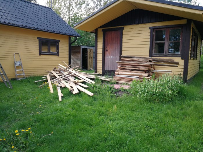 Virke som rensats från spik, staplat bredvid gult hus, med hög av virkesrester på gräs.