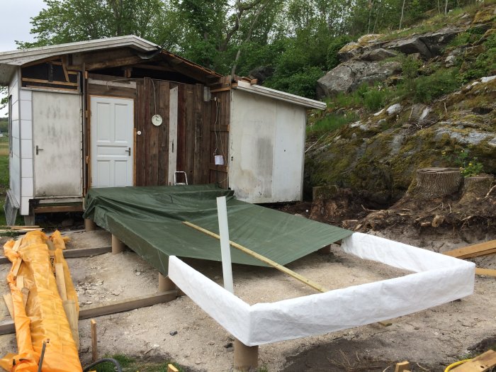 Pågående byggprojekt med synliga träramar och en grön presenning vid ett gammalt skjul med vit dörr.