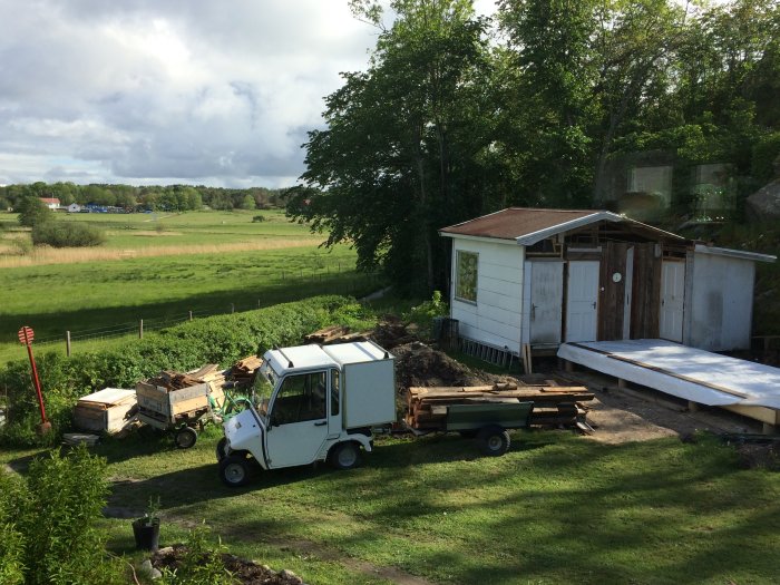 En liten lastbil full med rivningsmaterial bredvid ett halvfärdigt hus, omgivet av grönska och med byggmaterial framför.
