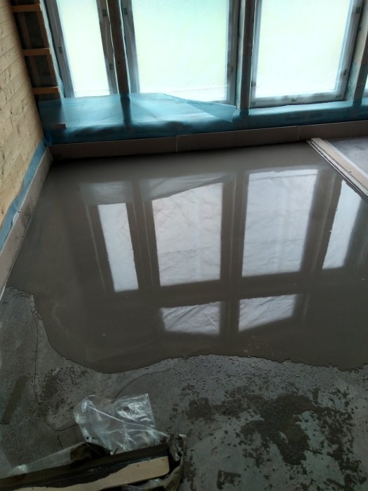 Nyutgjuten betonggolv reflekterar fönstret, med fukt och byggmaterial i förgrunden.
