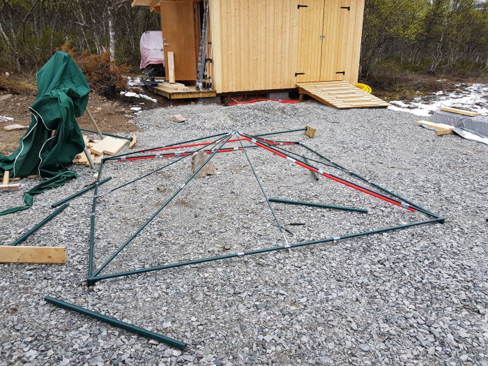 Söndrigt grön-rött partytältstomme ligger på en grusyta med en byggnad och skog i bakgrunden.