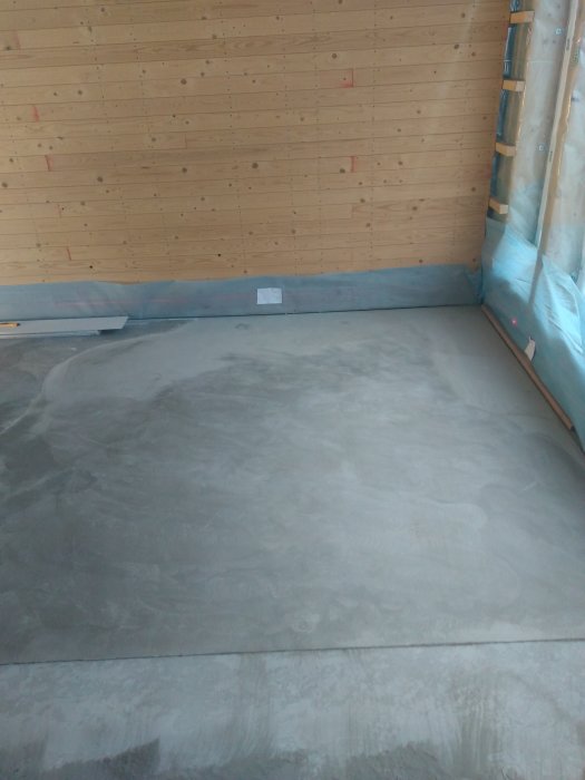 Nyligen slipad betonggolv med varierande jämnhet nära trävägg och plastöverdrag.