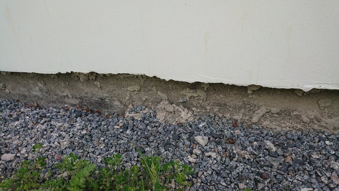 Skadad putsfasad i botten av ett hus med synligt fundament och grus, i behov av reparation.