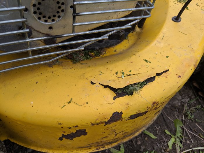 Närbild av ett sönderrostat gult klippdäck på en gräsklippare med gräsrester och synliga skador.