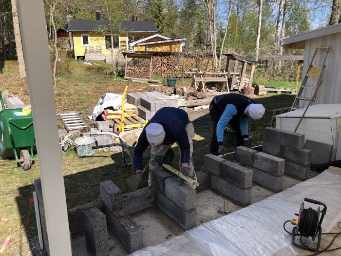 Två personer murar en konstruktion av lecablock utomhus med byggmaterial runtomkring.