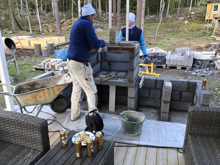 Två personer murar en konstruktion med lecablock utomhus, byggmaterial och ölburkar runtomkring.