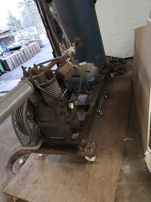 Gammal industrimaskin på en transportram med slangar och elmotor, placerad i ett garage.