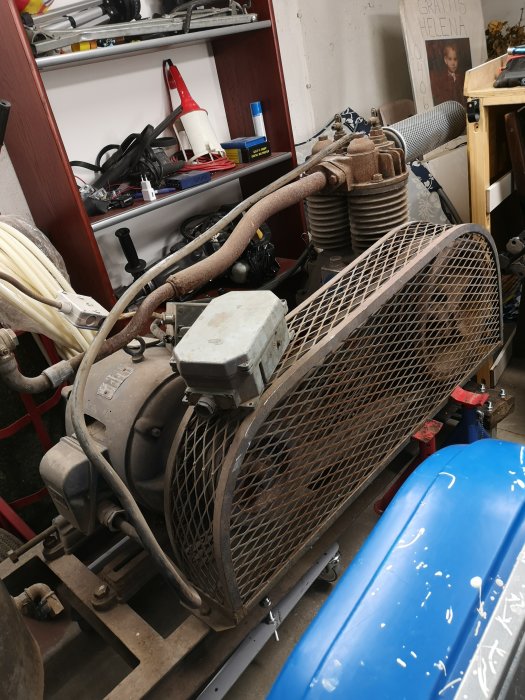 Äldre luftkompressor i ett garage, med slitage och anslutna luftslangar, står bredvid blå behållare.