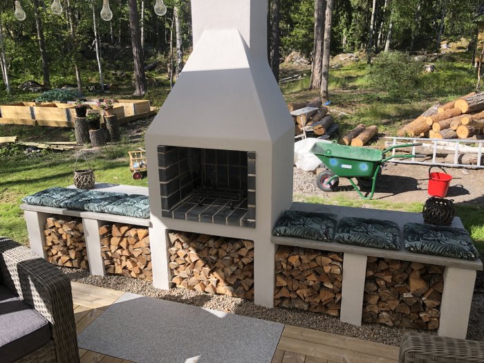 Nybyggd utomhusgrill med vedförvaring på en bakgård, omgiven av natur och trädgårdsredskap.