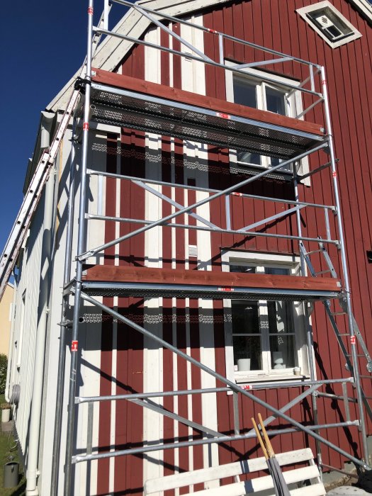 Byggnadsställning framför husvägg med nymålad fasad i rött och vitt, klart fram till fönstren.
