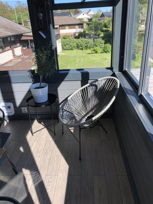 En nästan färdigbyggd altan med modern stol och växt framför glasräcke med utsikt över en grön trädgård.