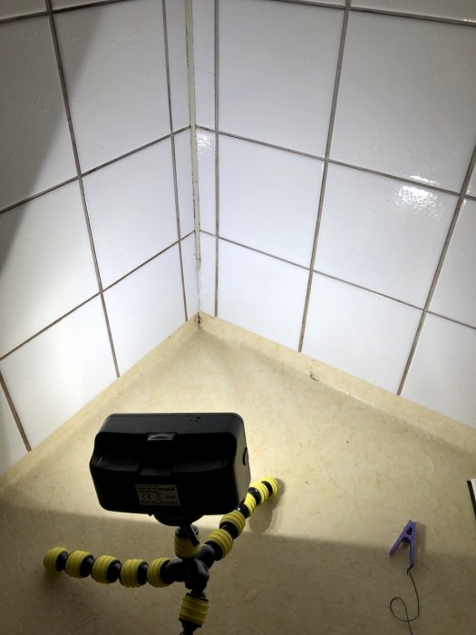 Hörn i ett badrum med skadat silikon, spruckna fogar mellan vita kakelplattor och en byggfläkt på golvet.