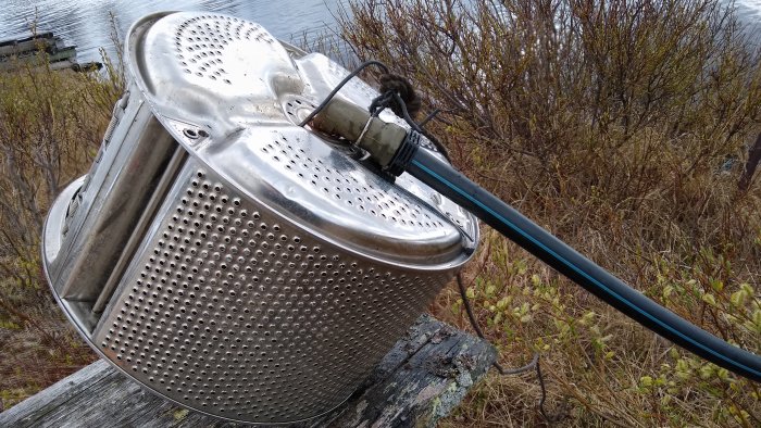 Ett uppochnervänt metallföremål, troligen en pumpautomat, med en ansluten svart slang, placerat utomhus med sjö och vegetation i bakgrunden.