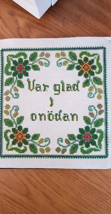 Broderi med texten "Var glad i onödan" omgivet av gröna blad och röda blommor på vit bakgrund.