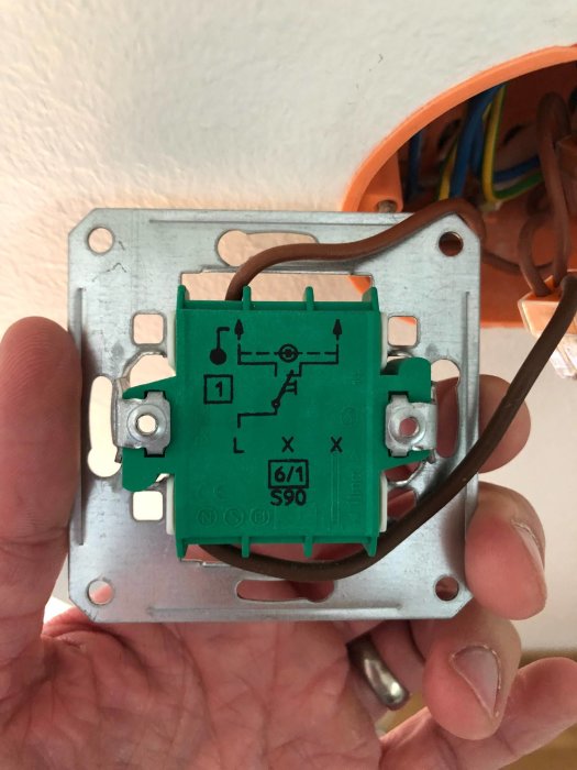 Hand håller en grön och grå ljusknapp monterad på vägg med synliga elektriska ledningar och anslutningar.