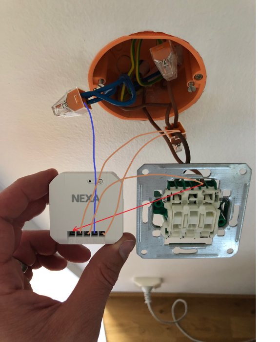 Hand håller Nexa strömbrytare med röd pil markerad, bredvid öppen installationsdosa med elektriska kablar och anslutningar.