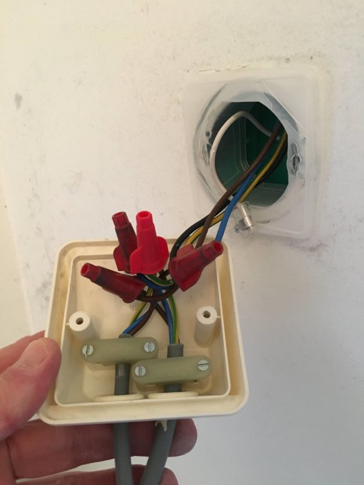 Hand håller en öppen elektrisk anslutningsdosa framför en vägg med utsatta kablar och ledningar, med en förseglad vit kabel.