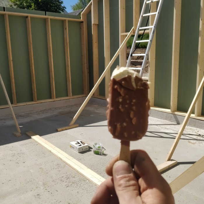 Hand som håller en glass framför en byggarbetsplats med trästommar och en stege i bakgrunden.