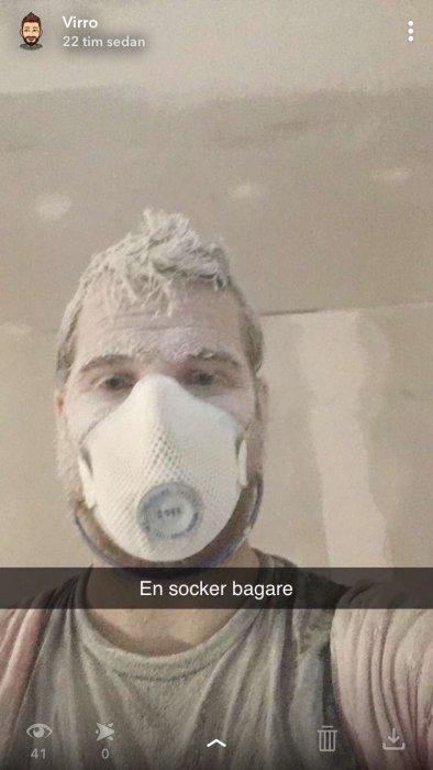 Person med skyddsmask täckt av spackeldamm framför nymålad vägg.