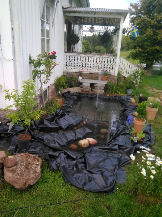 Nyanlagd regnträdgård med duk och stenar vid en vit husvägg, med en svensk flagga och planterade krukor runt omkring.