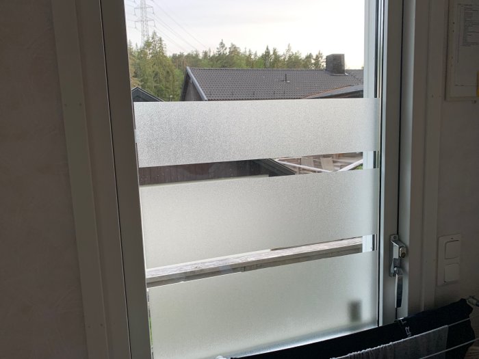Fönster med horisontella remsor av fönsterfolie för privatliv, med utsikt över trädtoppar.