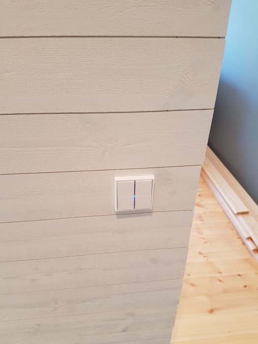 Nyinstallerad KNX-knapp på en texturerad vit vägg, med synlig trätrappa och golv i bakgrunden.