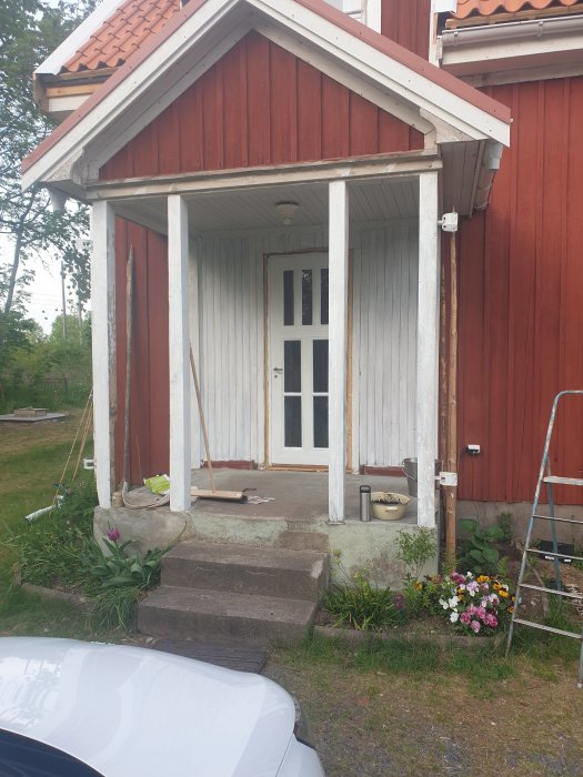 Renovations ongoing at a house façade with a white door framed by partially stripped wooden panelling under a porch, with construction items and plants visible.