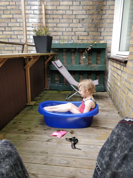 Barn i badkläder sitter i en blå barnpool på ett träaltandäck med husvägg i bakgrunden.