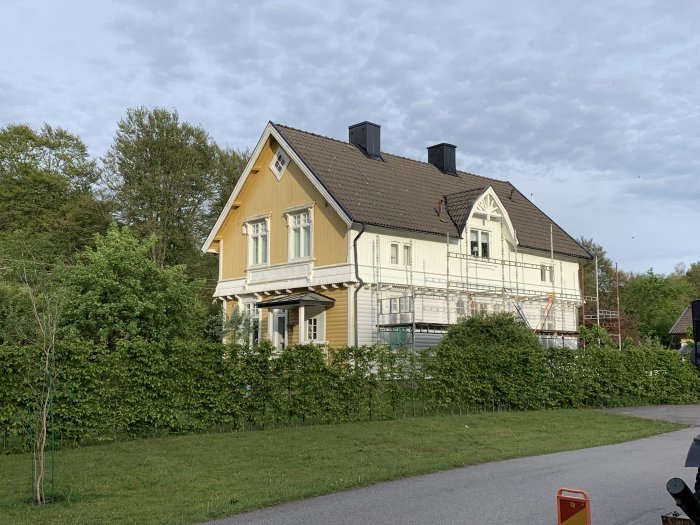 Gulmålat hus med byggnadsställningar under renovering omgiven av grönska.