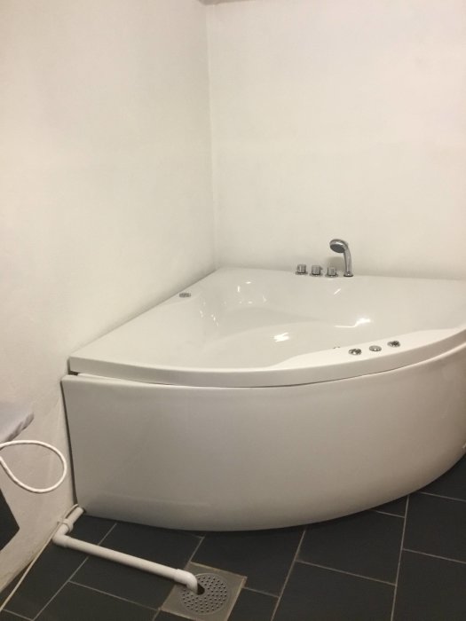 Ett hörnbadkar i ett pannrum med vita väggar och svart klinkergolv redo för renovering.