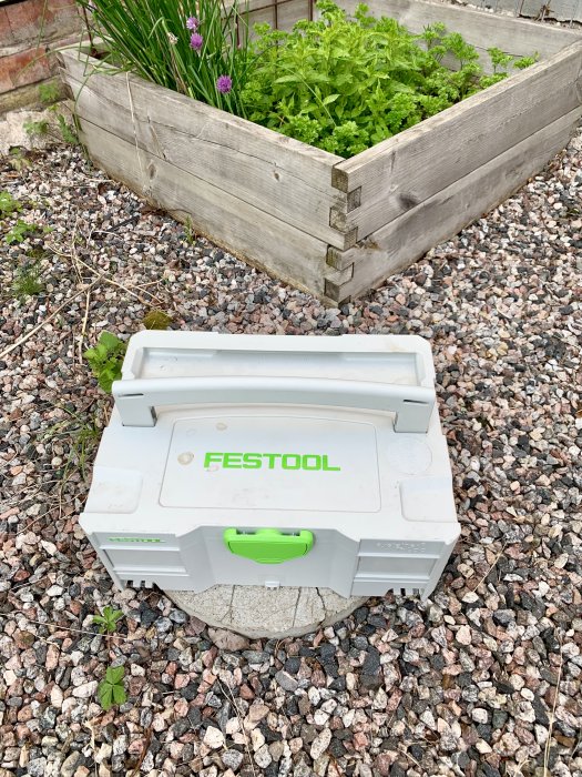 Festool verktygslåda framför en upphöjd träodlingslåda med gröna växter i en trädgård.