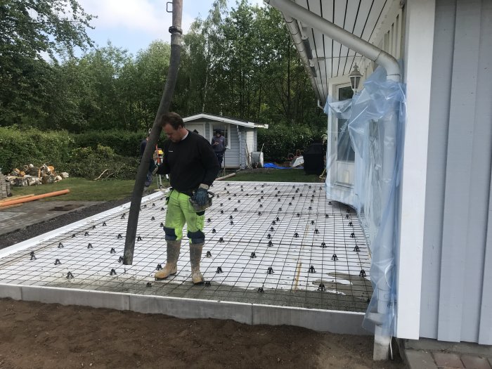 Person förbereder gjutning av betongplatta bredvid hus med armeringsnät och skyddad fasad.