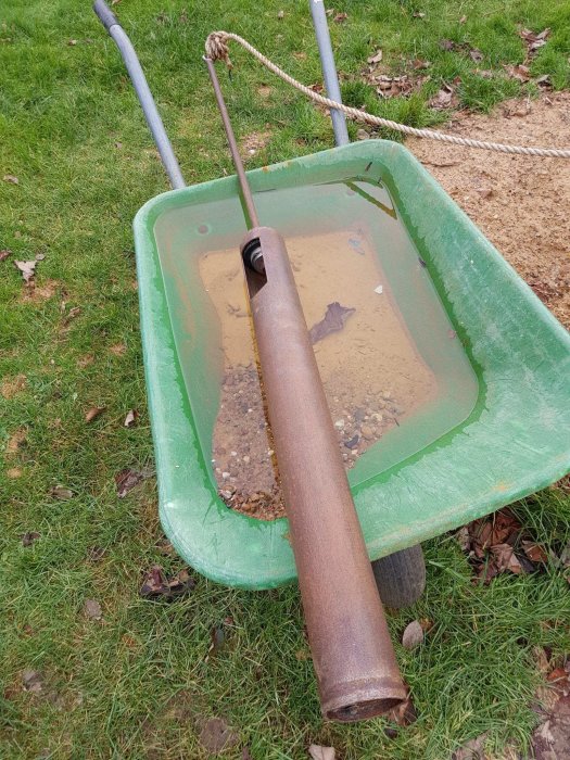 Grön skottkärra fylld med vatten och gravel, med en rostig metallrör i mitten.