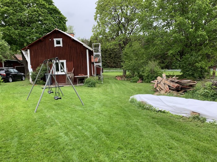 Röd stuga med vita knutar och stege, gungställning på grön gräsmatta, vedhög och ängsmark i bakgrunden.