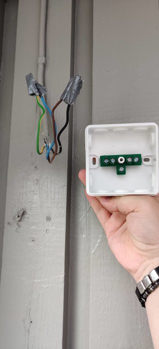 Hand håller en kopplingsdosa bredvid en vägg med fyra elektriska ledningar som sticker ut.