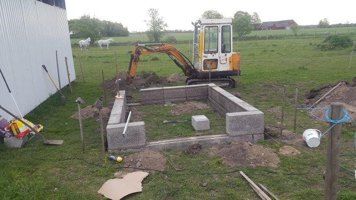 Grundkonstruktion av betongblock med armeringsstänger och grävmaskin på landsbygd.