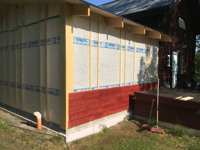 En nybyggd vägg med några grundmålade träpaneler och isoleringsmaterial mot en äldre byggnad.