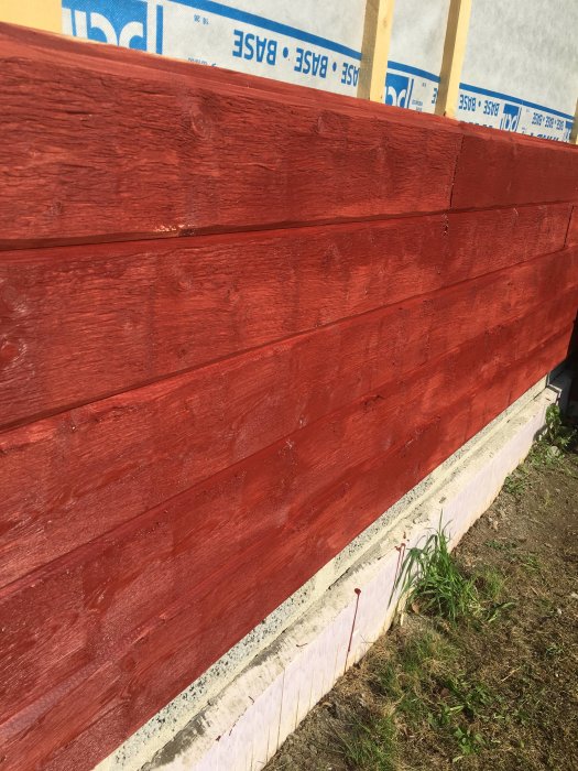 Nymålade röda panelbrädor på husvägg med synlig vindskyddsfolie och grundmur.