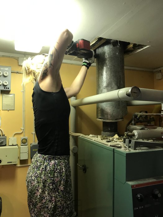 Kvinna arbetar med att kapade ett rör under innertaket med en elektrisk såg, omgiven av byggmaterial.