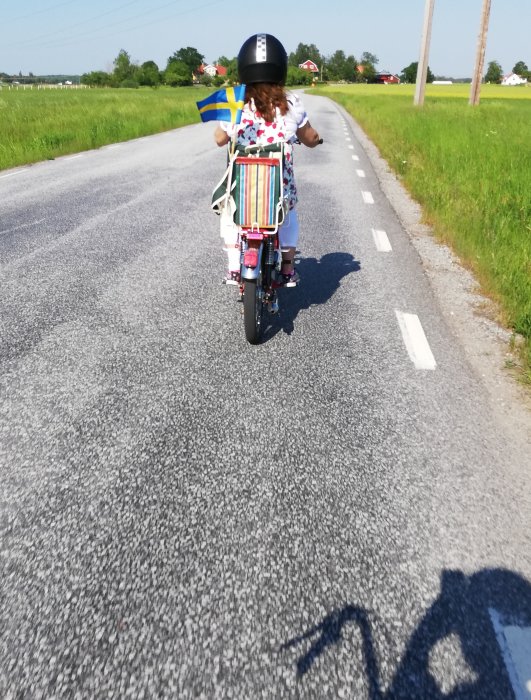 Barn i hjälm cyklar på landsväg och håller i en svensk flagga.