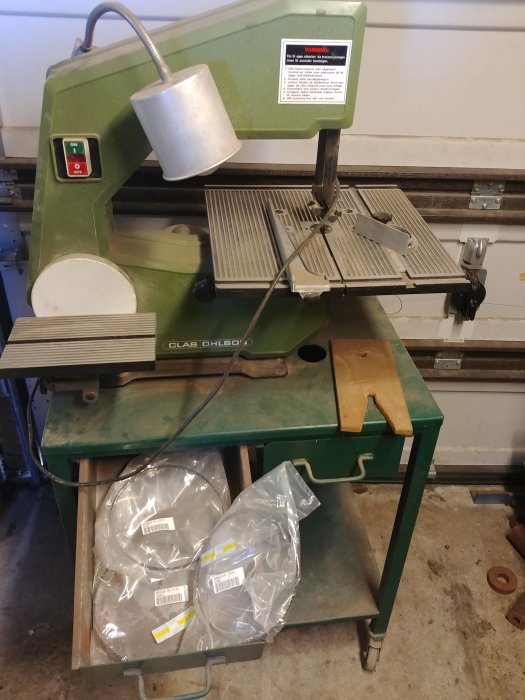 Grön bordsbandsåg av märket Clas Ohlson med arbetslampa och öppen låda med förpackade sågblad.