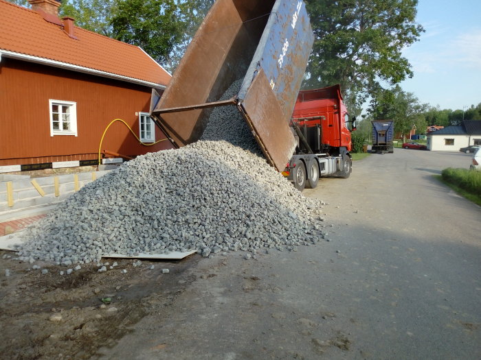 Lastbil tippande grus för paddning i byggprojekt, med en röd lastbil och ett hus i bakgrunden.